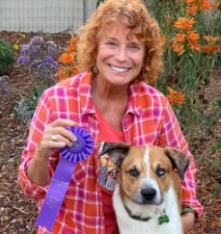 Woman with red hair and a pink/orange plain shirt smiling facing camera holding blue ribbon and a lab/border colllie mix dog